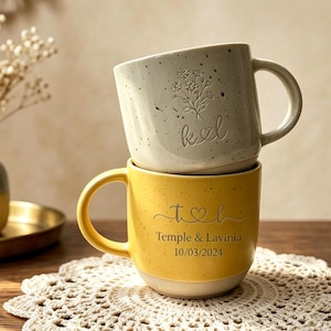 May include: Two stacked ceramic mugs with speckled finishes. The top mug is cream-colored with a floral engraving and the letters k and l. The bottom mug is yellow with the text Temple & Lavinia and the date 10/03/2024 printed below a heart icon.