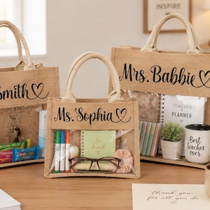 May include: Three tan jute tote bags with clear plastic windows and top handles. The bags are personalized with black script text reading Ms. Smith, Ms. Sophia, and Mrs. Babbie, each accompanied by a small heart icon on the right side.