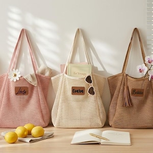 Könnte beinhalten: Drei Netztaschen in Rosa, Creme und Hellbraun, jeweils mit einem rechteckigen, braunen, lederartigen Aufnäher mit aufgedrucktem Namen. Die Taschen haben lange Schulterriemen und eine gewebte Struktur und sind nebeneinander angeordnet.