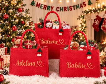 May include: Three red jute tote bags with sturdy handles, each personalized with gold cursive names Adelina, Salome, and Babbie followed by a heart symbol. Each bag features a small decorative tassel and is filled with various holiday items.