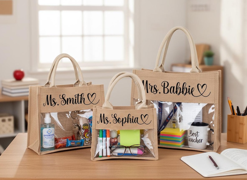 May include: Three rectangular jute tote bags with cream-colored handles and a clear plastic front panel. Each bag features black cursive text and a heart icon. The bags are personalized with the names Ms. Smith, Ms. Sophia, and Mrs. Babbie.