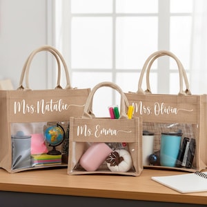 May include: Three tan burlap tote bags with clear plastic windows on the front. Each bag features white cursive text: Mrs. Natalie, Ms. Emma, and Mrs. Olivia. The bags have two top handles and are filled with various office and school supplies.