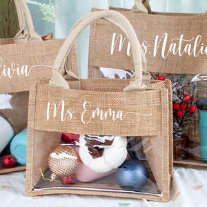 May include: Three rectangular tote bags made of tan burlap with two top handles and a clear plastic window on the front. Each bag features white cursive text reading Ms. Olivia, Ms. Emma, or Mrs. Natalie. The bags contain various items like tumblers.