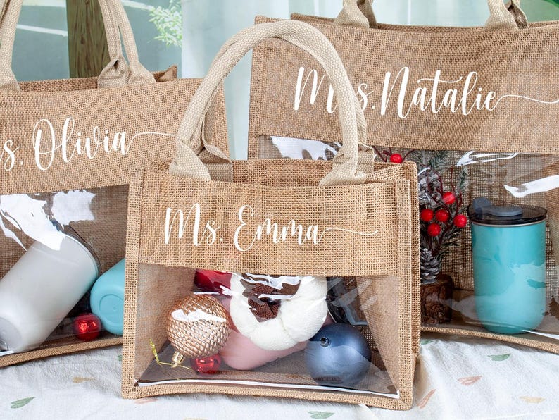 May include: Three rectangular tote bags made of tan burlap with two top handles and a clear plastic window on the front. Each bag features white cursive text reading Ms. Olivia, Ms. Emma, or Mrs. Natalie. The bags contain various items like tumblers.
