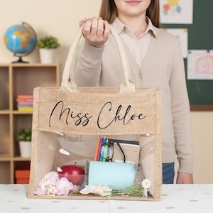 May include: A rectangular tote bag made of tan burlap with two cream-colored fabric handles. The bag features a clear plastic window on the front and the text "Miss Chloe" printed in black script. Inside are a red apple, a light blue cup, and books.