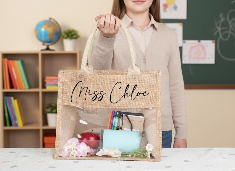 May include: A rectangular tote bag made of tan burlap with two cream-colored fabric handles. The bag features a clear plastic window on the front and the text "Miss Chloe" printed in black script. Inside are a red apple, a light blue cup, and books.