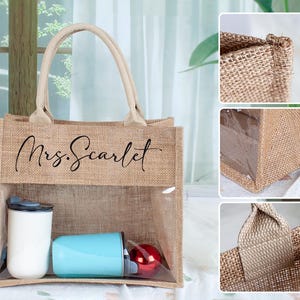 May include: A tan burlap tote bag with two beige handles and a clear plastic window on the front. The text Mrs. Scarlet is printed in black script on the upper burlap section. Inside the bag are a white tumbler, a light blue tumbler, and a red ornament.