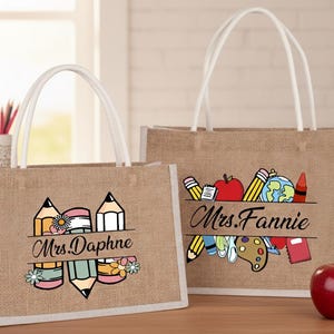 Puede incluir: Dos bolsas de mano de yute con asas blancas. Una bolsa presenta un diseño de lápiz y flores con el texto "Mrs. Daphne". La otra bolsa tiene un diseño de útiles escolares con el texto "Mrs. Fannie". Una manzana roja está cerca.