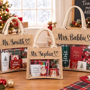 May include: Three tan burlap tote bags with clear windows, each personalized with names like Ms. Smith, Ms. Sophia, and Mrs. Babbie. The bags contain various items including tumblers with text like Teacher Fuel, notebooks, and small holiday-themed gift boxes.