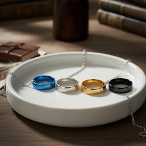 May include: Four rings on silver chains, presented on a white ceramic plate. The rings are blue, silver, gold, and black. The gold ring has engraved text. The background includes books and a small leather-bound book.