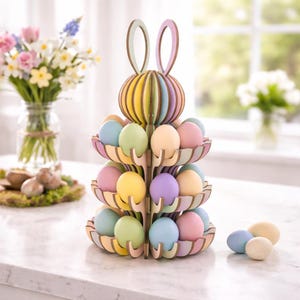 Puede incluir: Un soporte de madera para huevos de Pascua con forma de conejito, pintado en colores pastel. El soporte tiene tres niveles que sostienen huevos pintados en tonos azules, verdes, rosas, amarillos y morados. La cabeza y las orejas del conejito están en la parte superior.