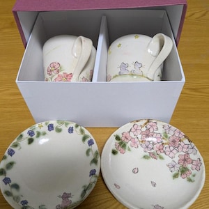 Pode incluir: Um conjunto de chá de cerâmica com duas canecas e dois pires. As canecas, numa caixa branca, apresentam motivos florais e de gatos. Os pires têm padrões florais e de folhas. O conjunto é pintado à mão com detalhes em rosa, roxo e verde.