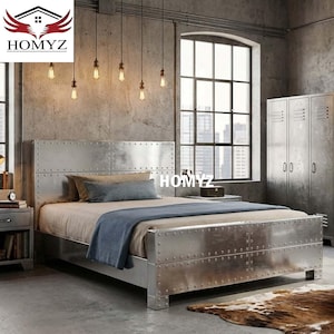 Puede incluir: Una cama de metal plateado con cabecero y piecero a juego. La cama tiene un diseño remachado y está en un dormitorio de estilo industrial. La habitación también cuenta con una gran ventana, luces colgantes y una alfombra de piel de vaca.