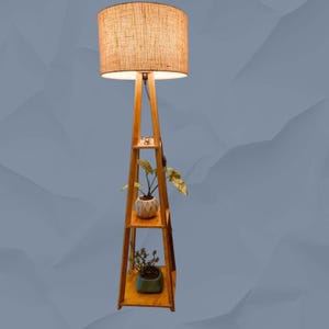 Op de afbeelding: Een houten vloerlamp met een beige linnen kap. De lamp heeft een ingebouwde plank met twee planken, elk met een potplant. De lamp is gemaakt van lichtgekleurd hout en heeft een eenvoudig, modern design.