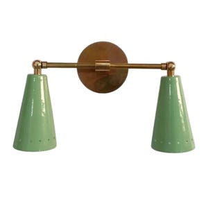 May include: A wall-mounted light fixture with two mint green cone-shaped shades. The fixture has a brass-colored circular backplate and a horizontal arm connecting the shades. Each shade has small, evenly spaced holes near the bottom.