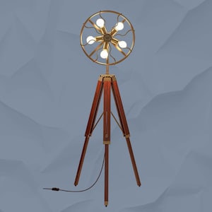 May include: A vintage-style floor lamp with a tripod base and a circular light fixture. The lamp features a dark wood tripod stand with brass accents. The light fixture has a gold-colored frame with six light bulbs. The lamp is plugged into a wall outlet.