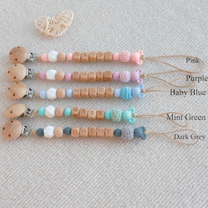 May include: A collection of baby pacifier clips in pink, purple, baby blue, mint green, and dark grey. Each clip features wooden beads with a name spelled out, along with decorative beads and a wooden clip. The names include Nando, Chris, Macca, Matt, and Chloe.