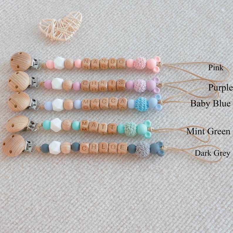May include: A collection of baby pacifier clips in pink, purple, baby blue, mint green, and dark grey. Each clip features wooden beads with a name spelled out, along with decorative beads and a wooden clip. The names include Nando, Chris, Macca, Matt, and Chloe.