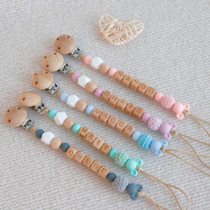 May include: Wooden baby pacifier clips with names spelled out in wooden letter beads. The clips feature various colors of silicone beads, wooden beads, and a small mouse-shaped silicone charm. A woven heart is in the background.