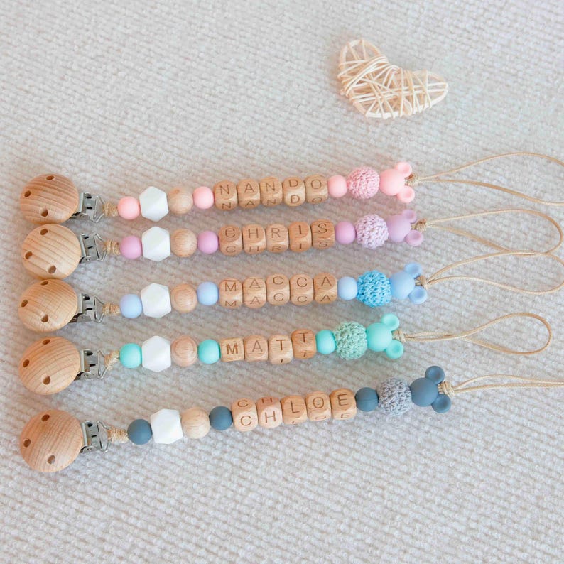 May include: Four personalized baby pacifier clips with wooden beads spelling out names. Each clip features a wooden clip, colorful beads, and a silicone animal accent. The names are Nando, Chris, Macca, and Matt. A small woven heart is in the background.