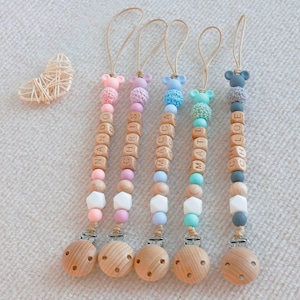 May include: Five personalized baby pacifier clips with wooden beads spelling out names. Each clip features a wooden base, colorful beads, and a small animal-shaped silicone top. The names are Nando, Chris, Macca, Matt, and Chloe.