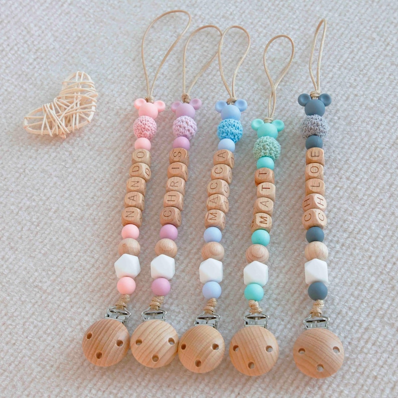 May include: Five personalized baby pacifier clips with wooden beads spelling out names. Each clip features a wooden base, colorful beads, and a small animal-shaped silicone top. The names are Nando, Chris, Macca, Matt, and Chloe.