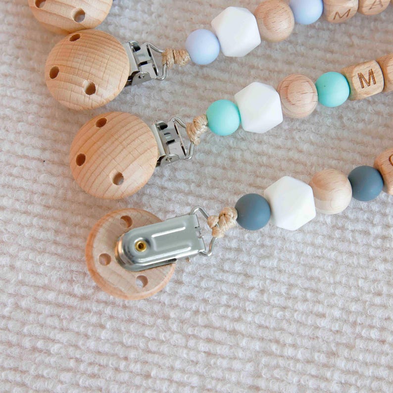 May include: Wooden baby pacifier clips with a natural wood finish, featuring a round clip and a beaded chain. The chains include light blue, white, and gray beads, along with wooden letter beads. The pacifier clips are designed for babies.