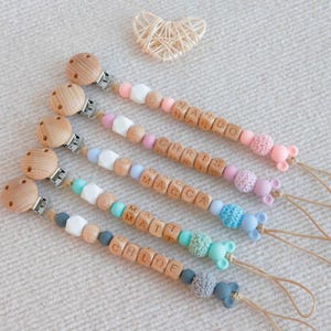 May include: Wooden baby pacifier clips with personalized names spelled out in wooden letter beads. The clips feature various colored beads, including pink, blue, and white, and are finished with a wooden clip and a small animal-shaped charm.
