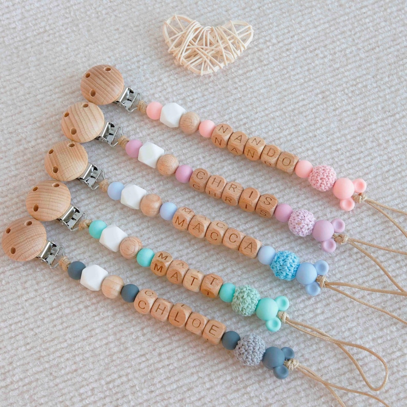 May include: Wooden baby pacifier clips with personalized names spelled out in wooden letter beads. The clips feature various colored beads, including pink, blue, and white, and are finished with a wooden clip and a small animal-shaped charm.