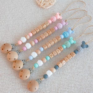 May include: Five personalized baby pacifier clips with wooden beads and letter blocks spelling out names. Each clip features a wooden round pacifier holder and a decorative end with a mouse-shaped design in various colors, including pink, blue, and gray.