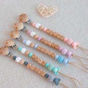May include: Five personalized baby pacifier clips with wooden beads spelling out names. Each clip features a wooden clip, colorful beads, and a silicone mouse-shaped end. The names include Nando, Chris, Macca, Matt, and Chloe.