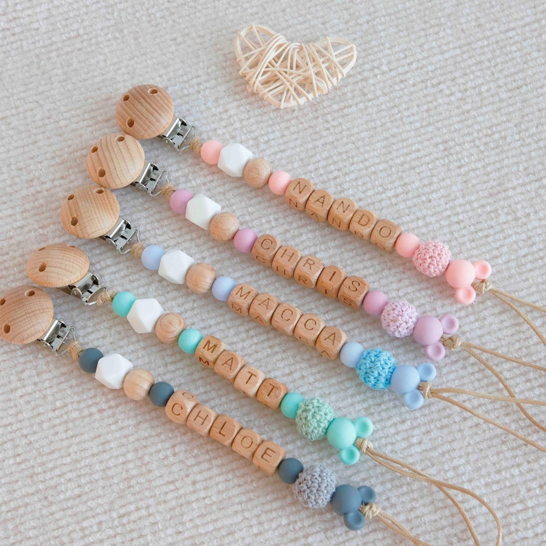 May include: Five personalized baby pacifier clips with wooden beads spelling out names. Each clip features a wooden clip, colorful beads, and a silicone mouse-shaped end. The names include Nando, Chris, Macca, Matt, and Chloe.
