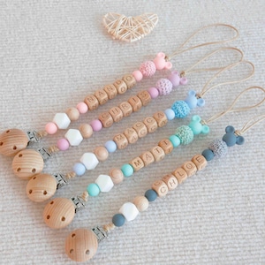 May include: A collection of personalized baby pacifier clips. Each clip features wooden beads spelling out a name, with colorful silicone beads and a wooden clip. The clips have a variety of pastel colors and decorative elements.