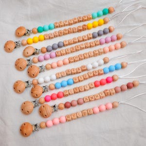 May include: A selection of personalised baby dummy clips. Each clip has a wooden clip, wooden letter beads spelling a name, and colourful silicone beads in various shades, including pink, blue, and yellow. Names include Bertrand, Valentina, Aurora, and others.