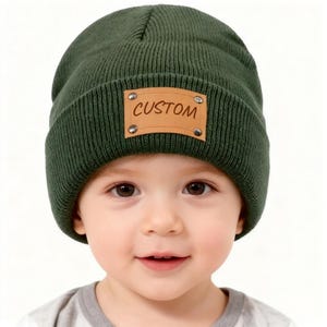 May include: A dark green knit beanie with a brown leather patch that reads "CUSTOM" in a handwritten style font. The patch is secured with four silver rivets. The beanie is shown on a child.