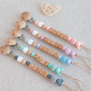 May include: Five personalized baby pacifier clips with wooden beads spelling out names. Each clip features a wooden clip, a variety of colorful beads, and a small animal-shaped silicone charm. The names are NANDO, CHRIS, MACCA, MATT, and CHLOE.