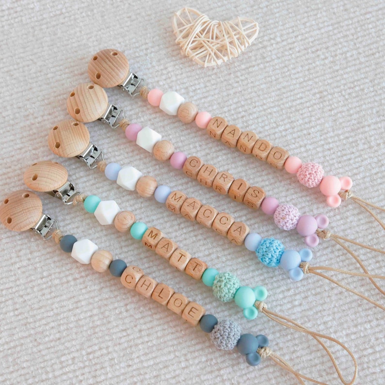 May include: Five personalized baby pacifier clips with wooden beads spelling out names. Each clip features a wooden clip, a variety of colorful beads, and a small animal-shaped silicone charm. The names are NANDO, CHRIS, MACCA, MATT, and CHLOE.