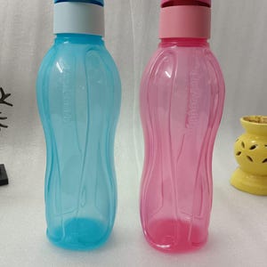 Könnte beinhalten: Zwei Wasserflaschen aus Kunststoff, eine hellblaue und eine rosa, mit farblich passenden Deckeln. Die Flaschen haben ein geschwungenes, ergonomisches Design und der Markenname ist auf der Seite aufgedruckt.
