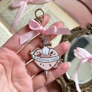 Peut inclure: Un porte-clés rose et doré avec un pendentif en forme de cœur. Le pendentif représente une épée, des roses et l'inscription "Romantasy reader" sur une bannière. Un ruban rose est attaché. En arrière-plan, un livre, des fleurs et un miroir.