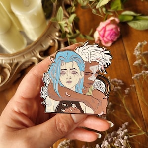 May include: A metal pin depicting two figures in an embrace, framed by white roses. One figure has blue hair, the other has white hair. The pin is held in a hand, with a blurred background of candles, flowers, and foliage.