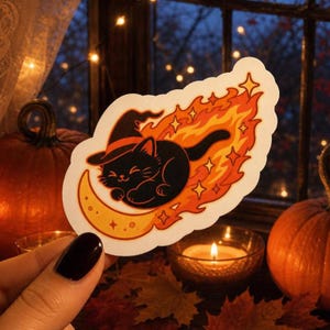 May include: A die-cut sticker featuring a black cat wearing a pointed witch hat, curled up on a yellow crescent moon. The cat is surrounded by stylized orange and red flames with small four-pointed star accents against a white background.