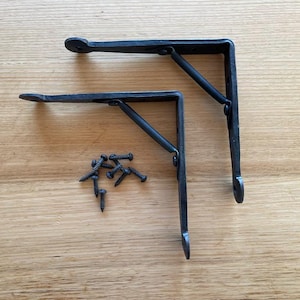 May include: Two black, L-shaped metal shelf brackets with a decorative design, and a small pile of black screws. The brackets are on a light brown wooden surface. The brackets are designed to support shelves.