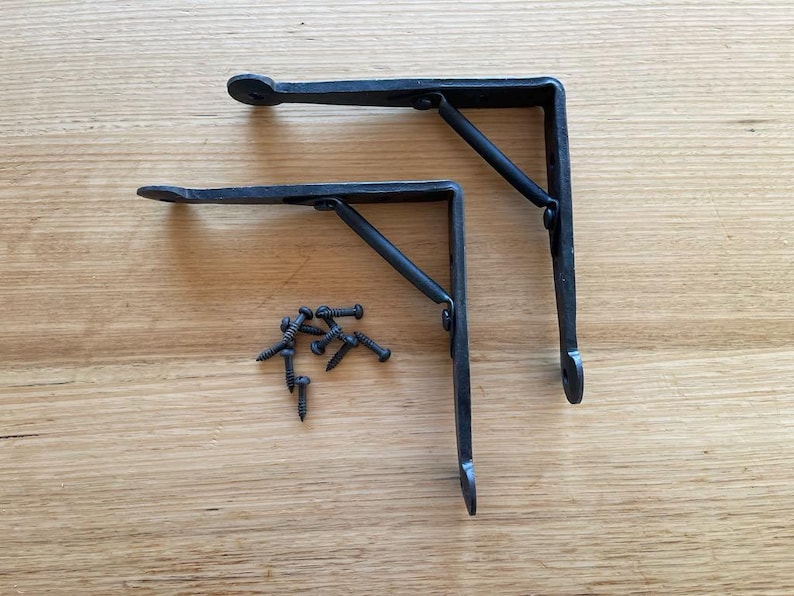 May include: Two black, L-shaped metal shelf brackets with a decorative design, and a small pile of black screws. The brackets are on a light brown wooden surface. The brackets are designed to support shelves.