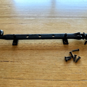 May include: Black wrought iron window or door latch with mounting brackets and screws. The latch has a decorative curved end and a pivoting arm to secure a window or door. The screws are black.