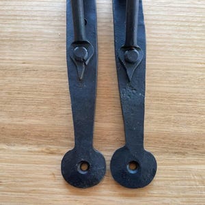 May include: Two black, wrought iron door pulls with a handle and an elongated backplate. The backplates have a circular hole at the bottom for mounting. Several screws are visible on the left. The door pulls are on a light brown wooden surface.
