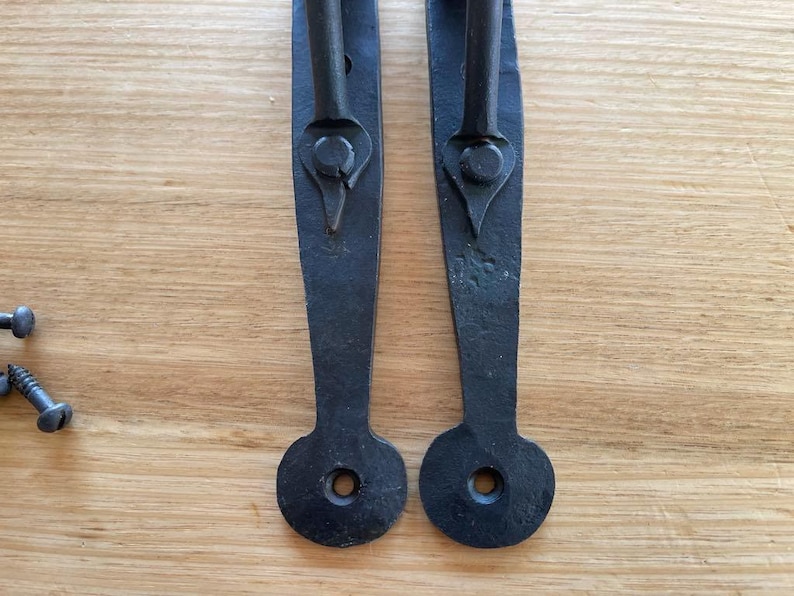 May include: Two black, wrought iron door pulls with a handle and an elongated backplate. The backplates have a circular hole at the bottom for mounting. Several screws are visible on the left. The door pulls are on a light brown wooden surface.