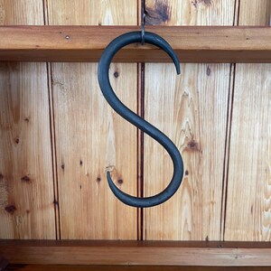May include: A dark grey, S-shaped metal hook suspended from a wooden shelf. The hook is set against a backdrop of vertical wooden planks. The hook's design is simple and functional, suitable for hanging items.