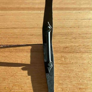 May include: A black, hand-forged iron door latch. The latch has a circular end with a hole, a long, curved body, and a central bar secured with rivets. The metal has a textured, rustic appearance, suggesting a vintage or antique style.