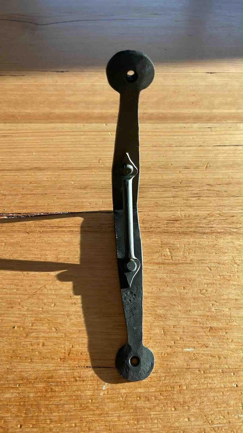 May include: A black, hand-forged iron door latch. The latch has a circular end with a hole, a long, curved body, and a central bar secured with rivets. The metal has a textured, rustic appearance, suggesting a vintage or antique style.