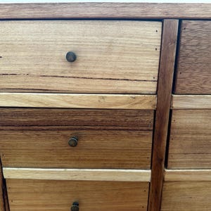 May include: Close-up of a wooden cabinet with multiple drawers. The drawers are made of various shades of wood, from light tan to dark brown, and have small, round, bronze-coloured knobs. The cabinet's frame is a darker brown, and the wood grain is visible.